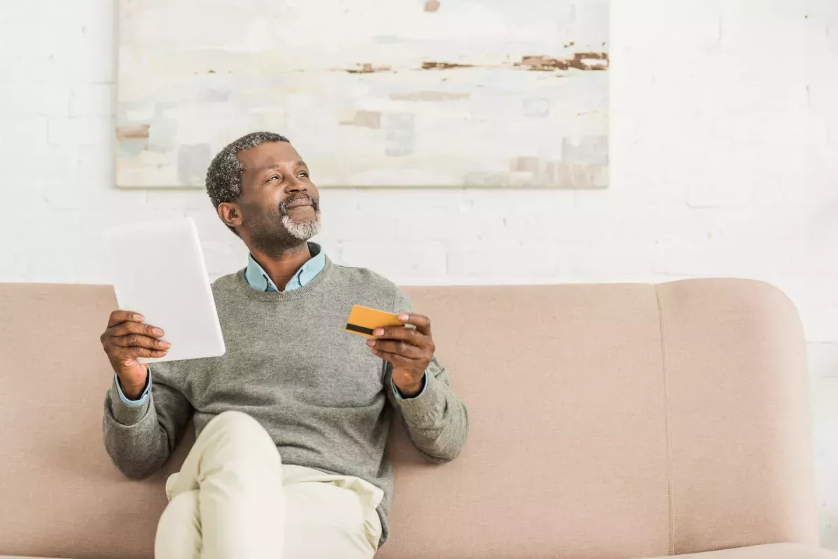 AdobeStock_300378721.jpeg smiling man sitting on couch with credit card and phone