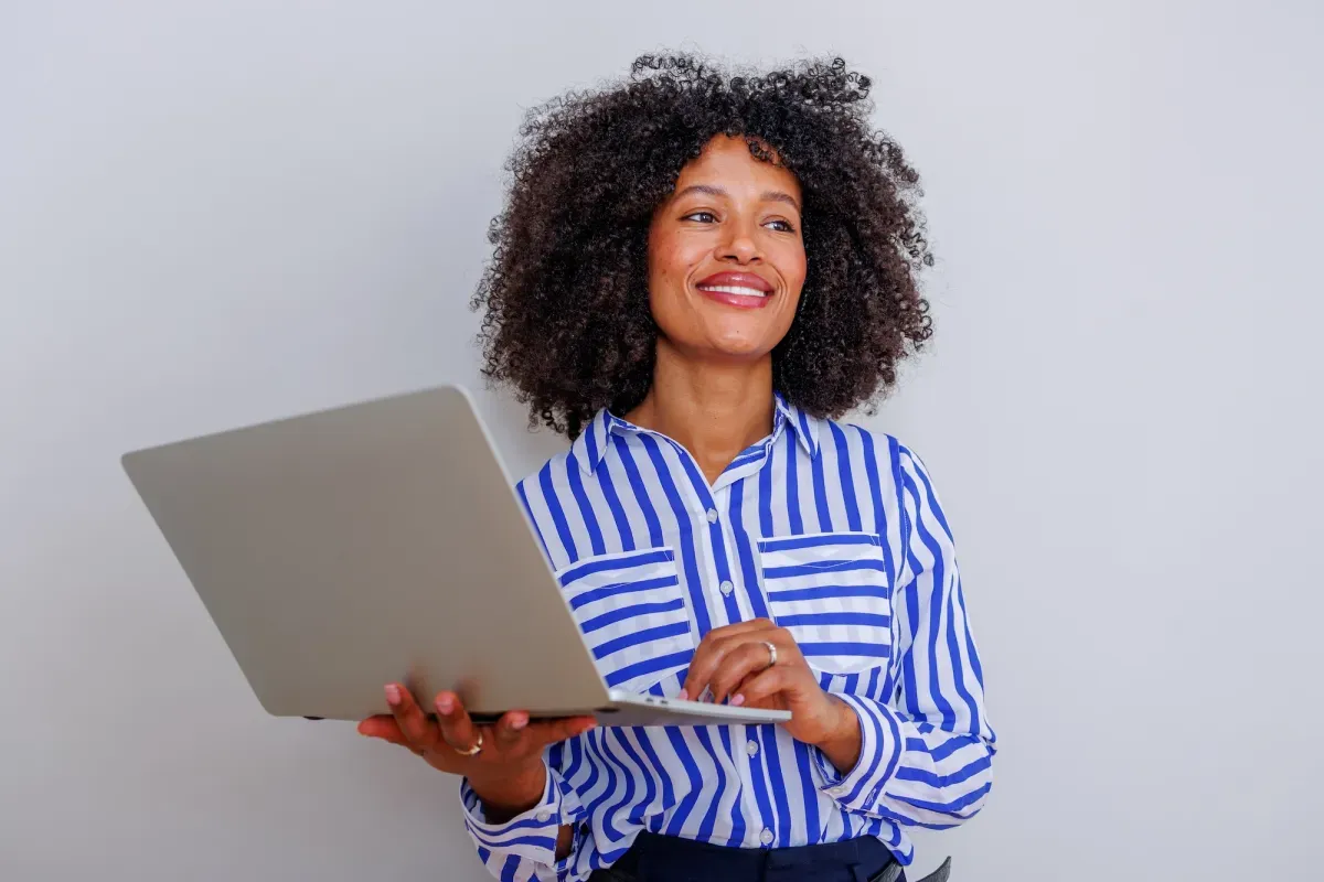 Smiling woman with curly hair in blue striped shirt holding laptop against gray background Smiling woman with curly hair in blue striped shirt holding laptop against gray background