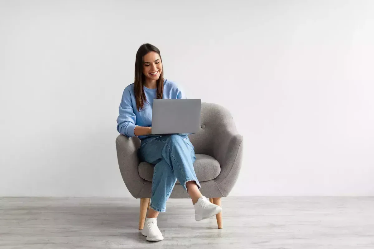 Young woman in blue outfit sitting in gray armchair, smiling while using a laptop. Young woman in blue outfit sitting in gray armchair, smiling while using a laptop.