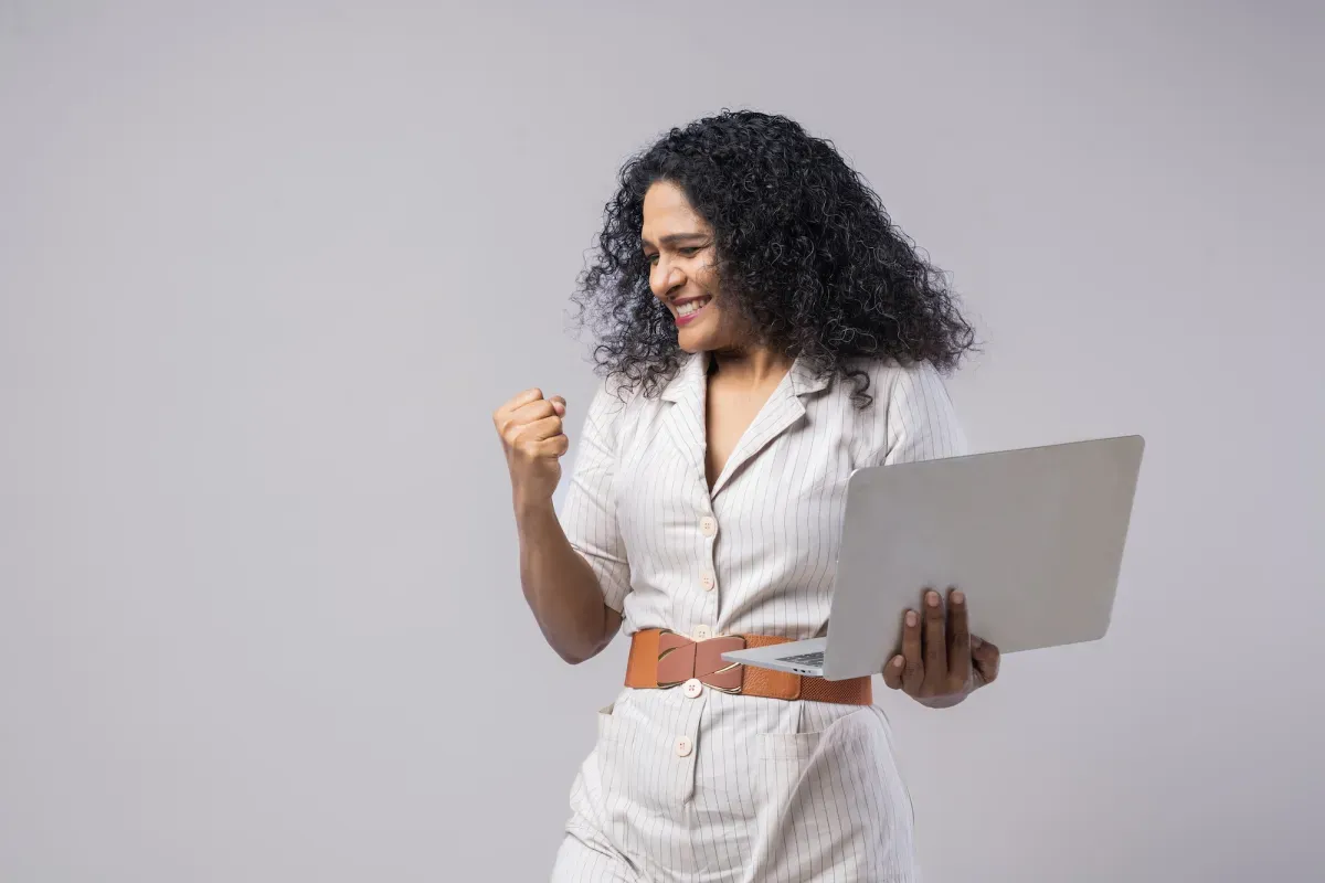 Woman in white outfit celebrating with raised fist while holding a laptop.