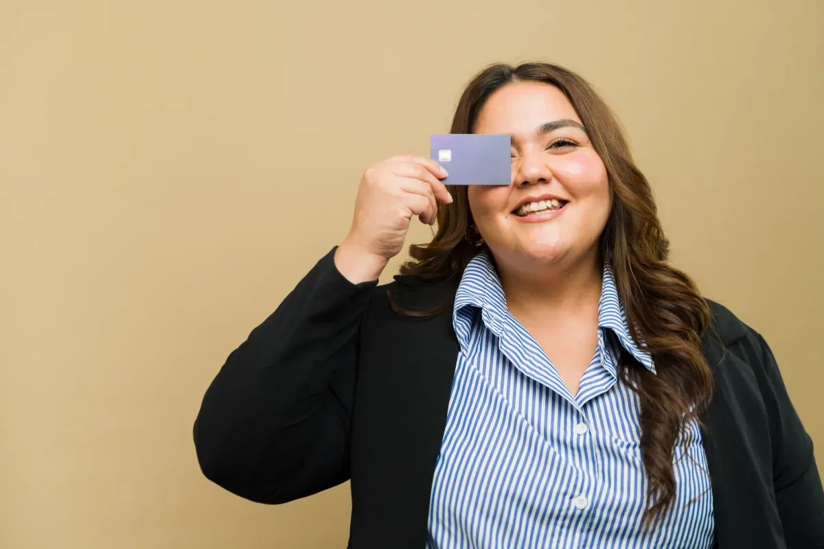 Smiling woman in a blazer holding a credit card over one eye against a beige background.