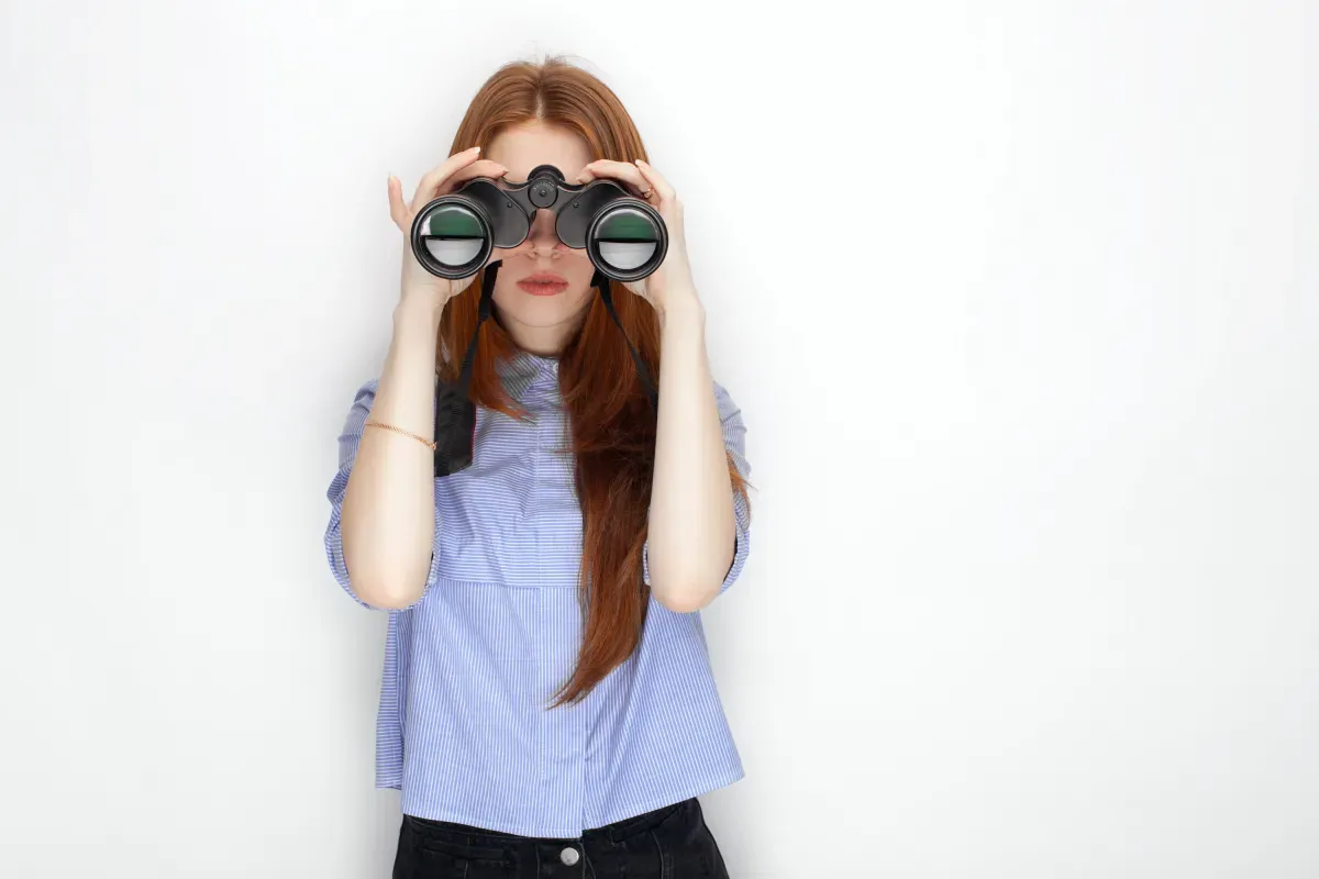 Red-haired woman in a light blue shirt looking through black binoculars against a plain white background.
