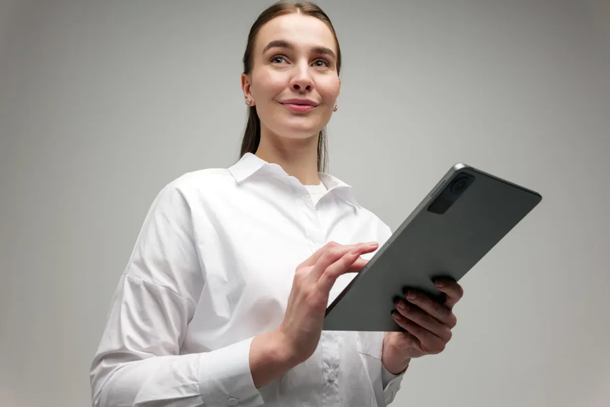 Woman in a white button-up shirt smiling slightly while using a tablet against a plain gray background. Woman in a white button-up shirt smiling slightly while using a tablet against a plain gray background.