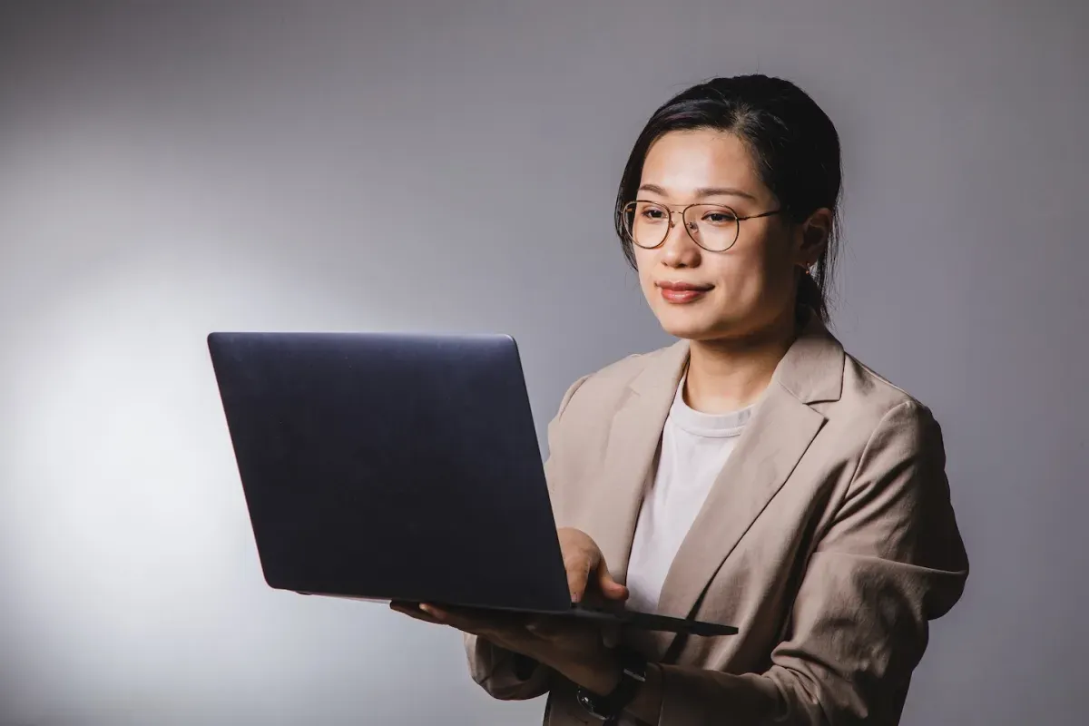Smiling woman with glasses and a beige blazer looks at an open laptop against a plain gray background. Smiling woman with glasses and a beige blazer looks at an open laptop against a plain gray background.