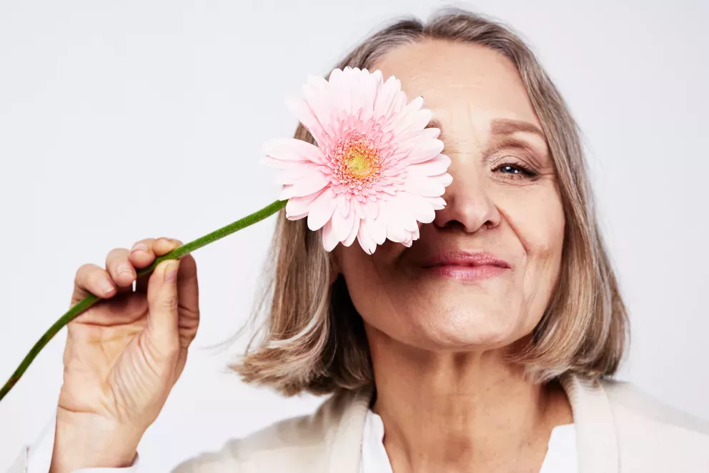 smiling senior woman holding pink daisy over one eye