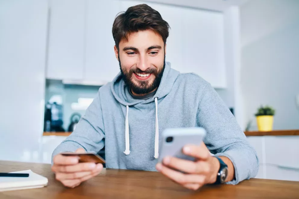 smiling young man sitting at home counter and paying online with credit card and smartphone