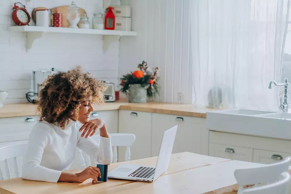 smiling woman sitting at kitchen table with laptop and credit card