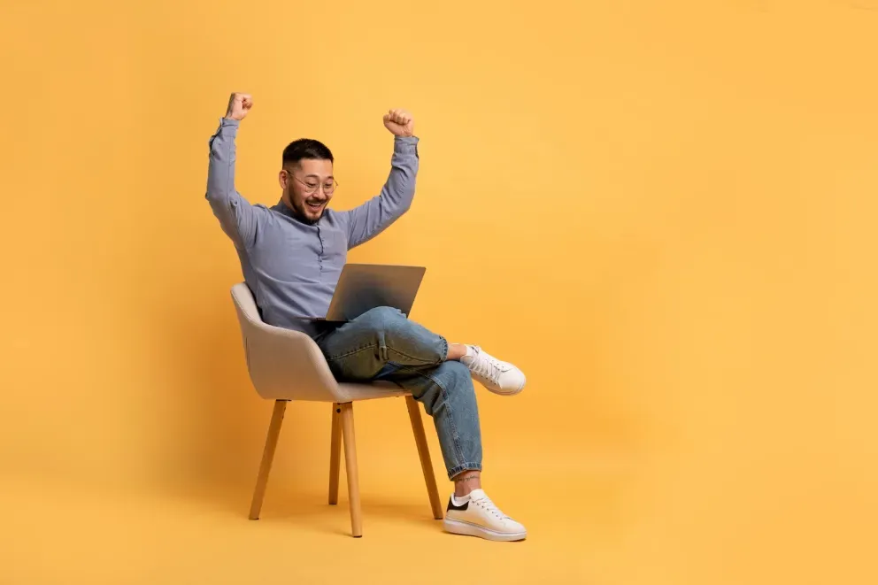 excited man sitting in chair with laptop throwing arms up in celebration