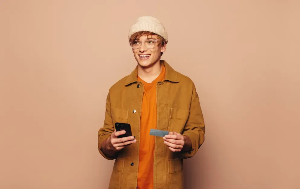 Young man in burnt orange jacket and beanie holding his phone and credit card smiling. Young man in burnt orange jacket and beanie holding his phone and credit card smiling.