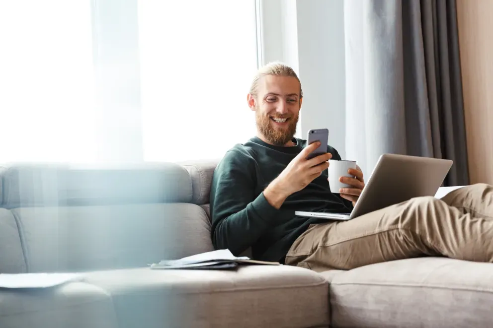 Smiling man on couch with coffee phone and laptop