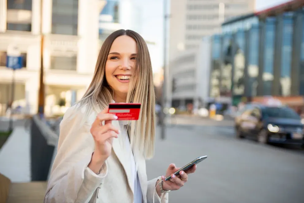 Woman in white blazer holding credit card and smartphone on city street
