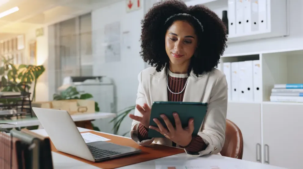 Black professional woman with curly hair reviewing a tablet at her office desk.