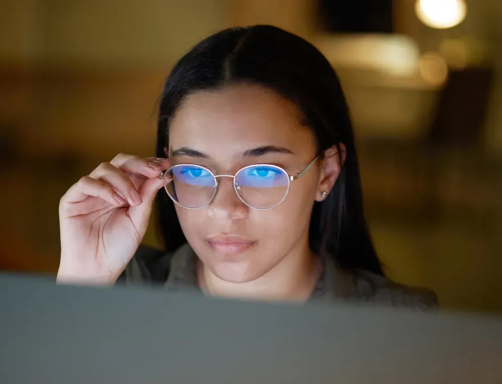 Young woman peering over glasses at a computer screen in a dimly lit office.