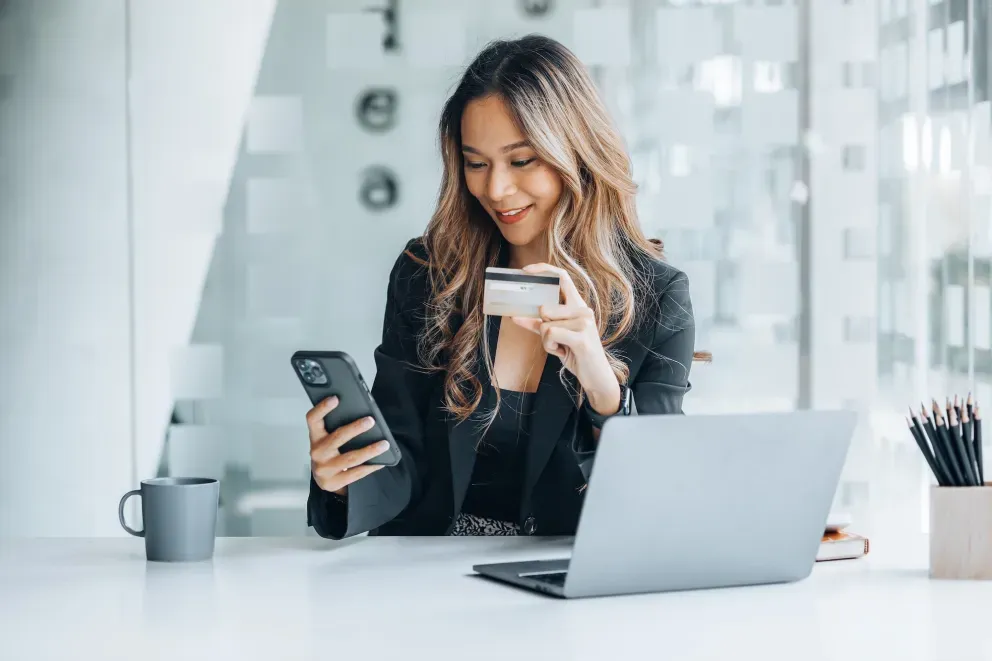 Young woman smiling, holding a credit card and smartphone while sitting at a laptop.