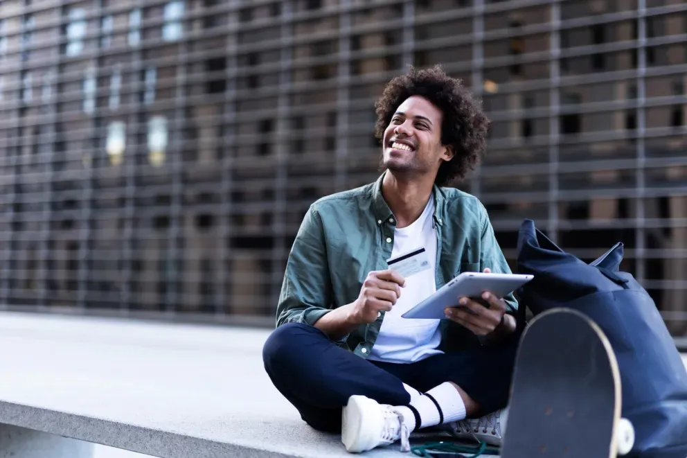 Young man with curly hair sitting outdoors, laughing while holding a tablet and pen.
