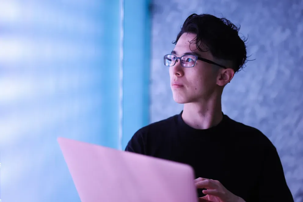 Young man with glasses holding a laptop, illuminated by blue and purple neon light.