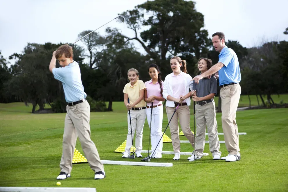 Young boy swinging a golf club on a green course while an instructor and children watch. Young boy swinging a golf club on a green course while an instructor and children watch.