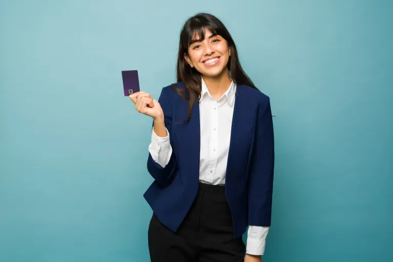 Young woman in a navy blazer smiling while holding a credit card against a teal background.