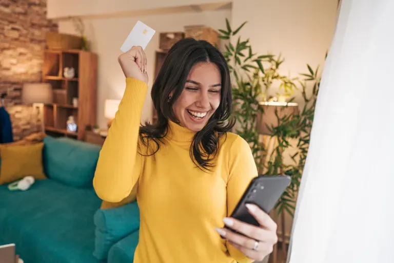 Young woman in yellow sweater celebrating, holding a credit card and smartphone indoors.