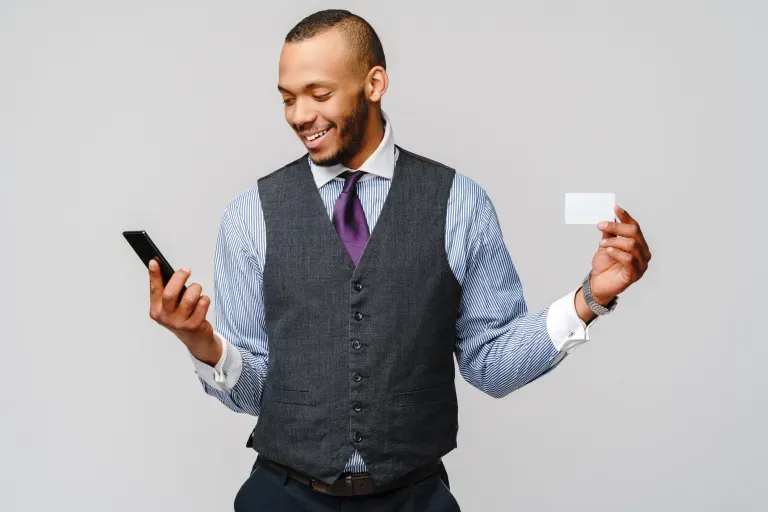 Smiling man in vest holding a smartphone and credit card against a gray background.