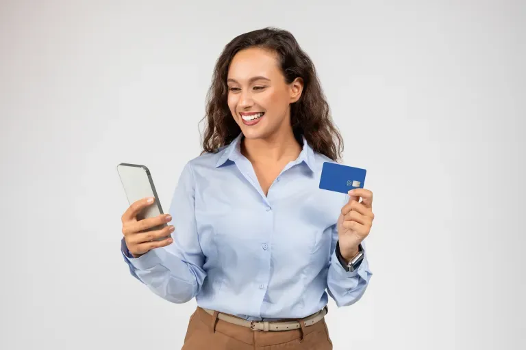Smiling young woman holding a smartphone and blue credit card against a light gray background.