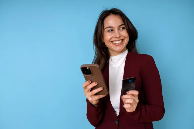 Young woman in maroon blazer and white turtleneck smiling ,holding a brown covered phone against a bright blue background.