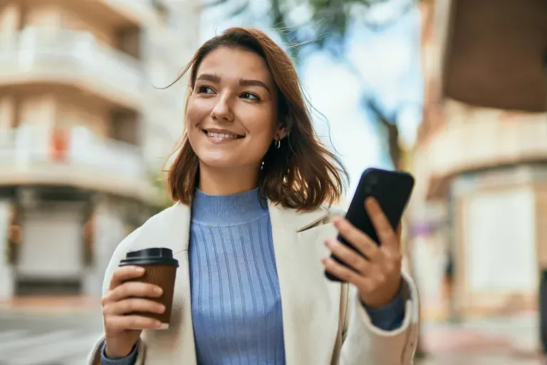 Young woman smiling outdoors, holding a coffee cup and smartphone on a city street.