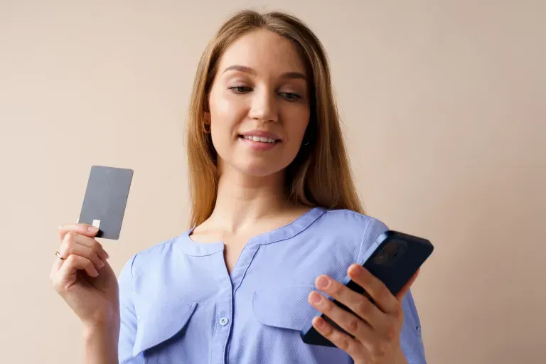 Young woman in a blue blouse smiling,holding a credit card and phone against a beige background.