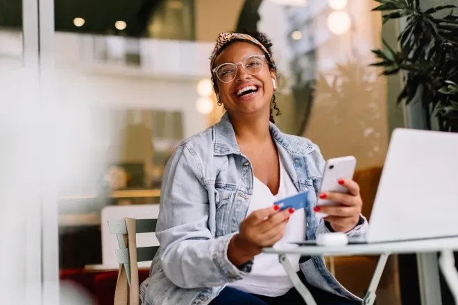 Young woman laughing while holding a smartphone and credit card at a café table.