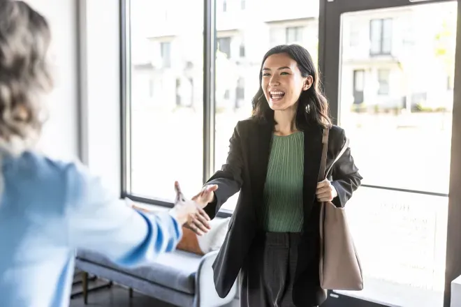 Two women shaking hands in a bright office entrance, one smiling warmly in a black blazer.