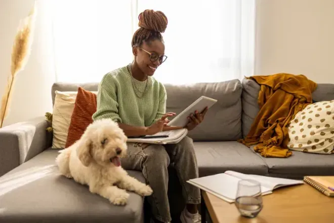Young woman reading a book on a gray couch beside a fluffy white dog in a cozy living room. Young woman reading a book on a gray couch beside a fluffy white dog in a cozy living room.