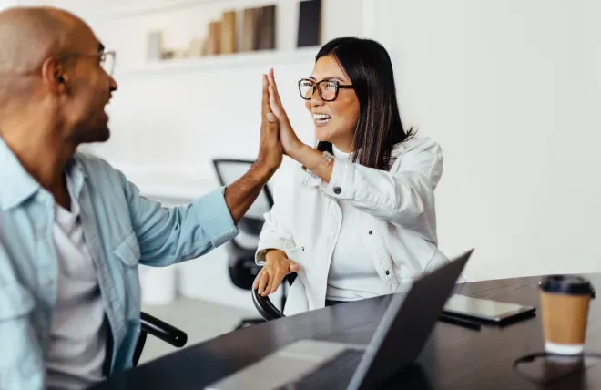 Two coworkers high-fiving and smiling at each other in a bright office setting. Two coworkers high-fiving and smiling at each other in a bright office setting.