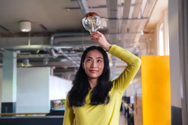 Woman in yellow sweater holding a large lightbulb above her head, smiling confidently indoors. Woman in yellow sweater holding a large lightbulb above her head, smiling confidently indoors.