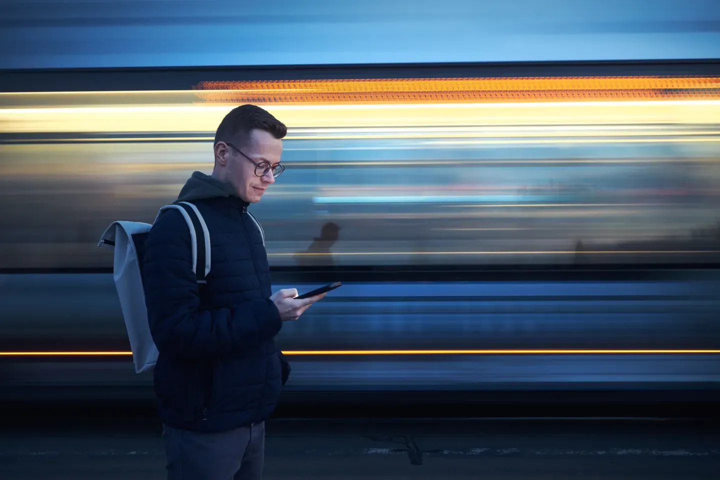 Man in dark jacket with phone as blue train speeds past in motion blur