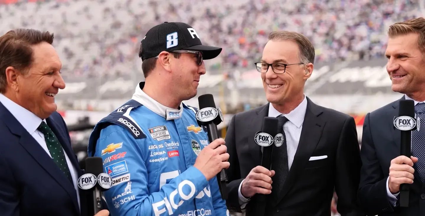 NASCAR driver in blue racing suit speaking with officials and team members in garage area