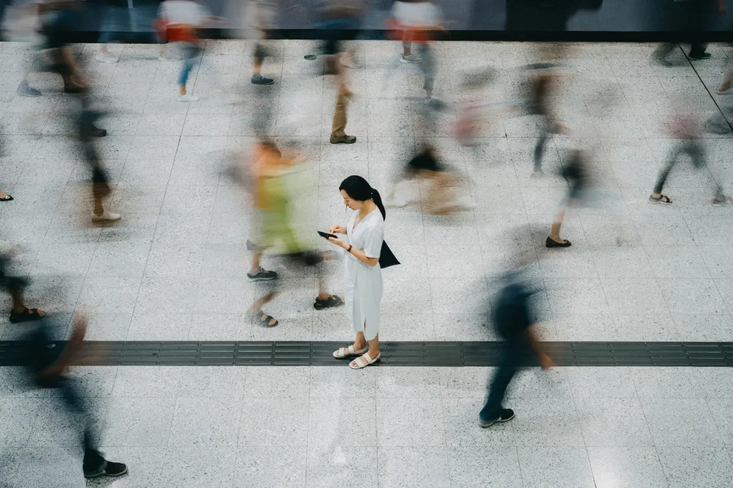 Woman standing still looking at phone amid blurred crowd of rushing pedestrians, aerial view.