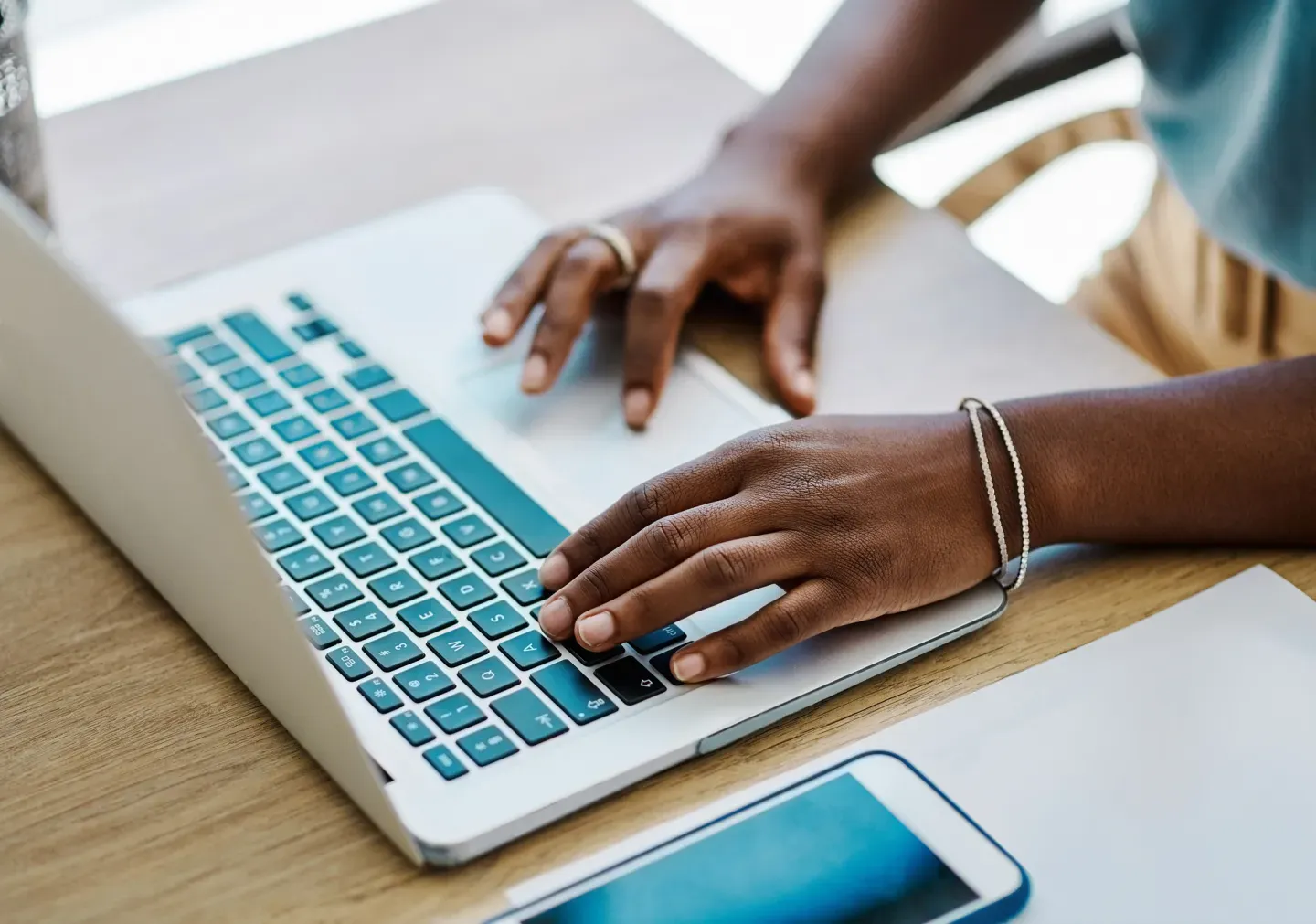 Person typing on a silver laptop, with a smartphone beside it on a wooden desk. Person typing on a silver laptop, with a smartphone beside it on a wooden desk.