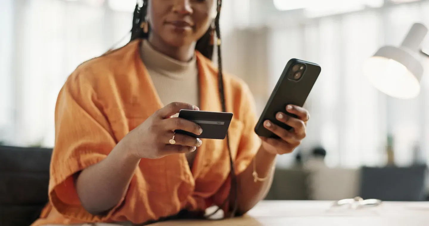 Woman in an orange cardigan holds a credit card in one hand and a smartphone in the other, seated indoors. Woman in an orange cardigan holds a credit card in one hand and a smartphone in the other, seated indoors.