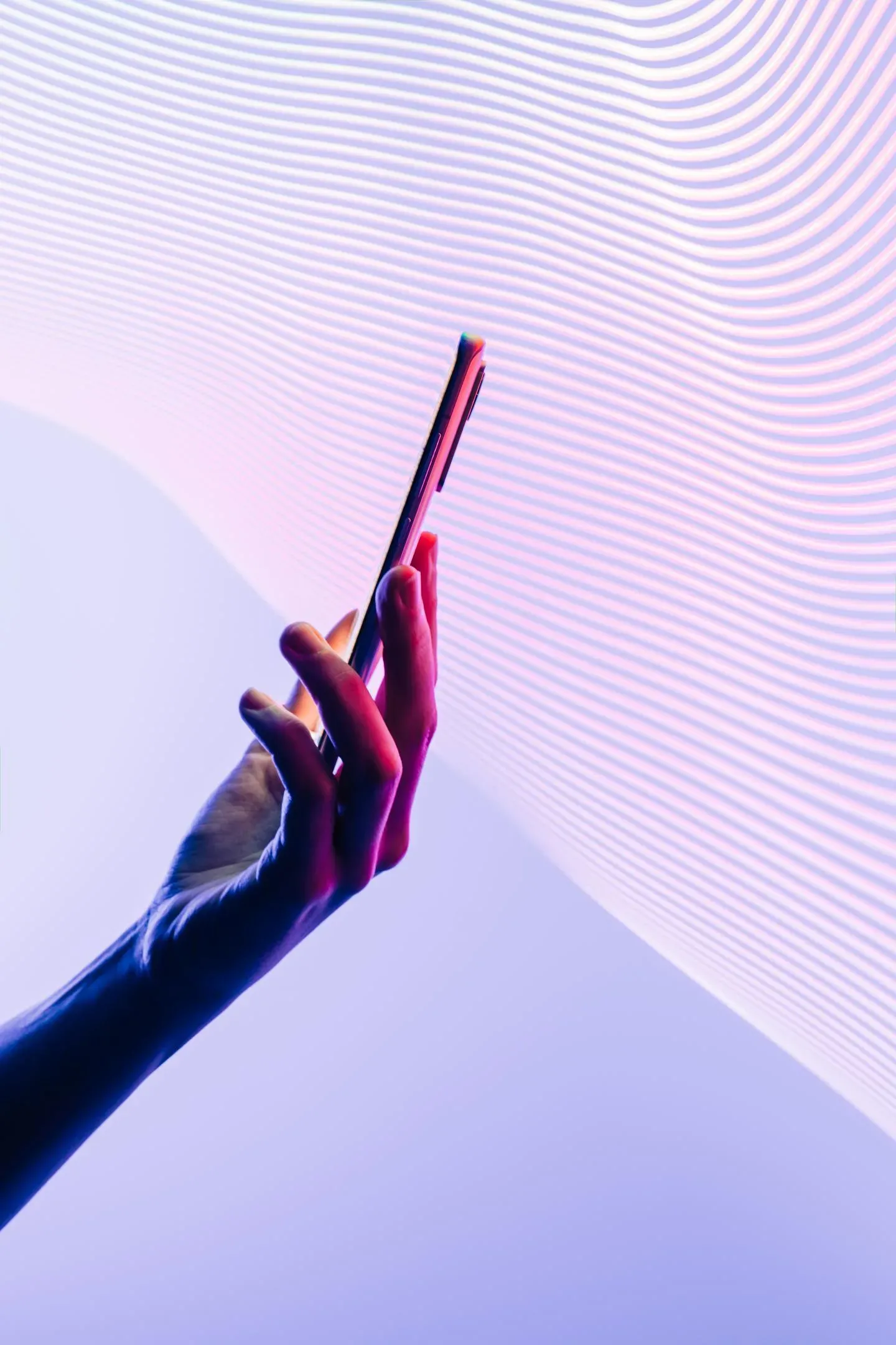 Hand lit in pink and blue holding a smartphone against a purple background with flowing white wave lines. Hand lit in pink and blue holding a smartphone against a purple background with flowing white wave lines.