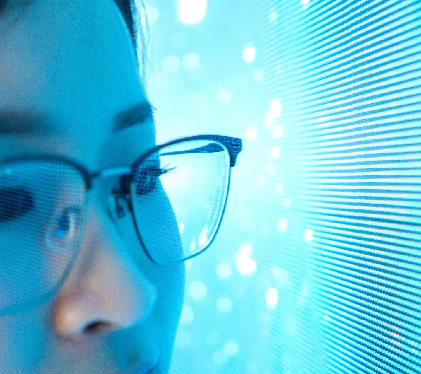 Close-up of a person in glasses, lit in blue, gazing at a glowing screen of fine lines and light dots. Close-up of a person in glasses, lit in blue, gazing at a glowing screen of fine lines and light dots.