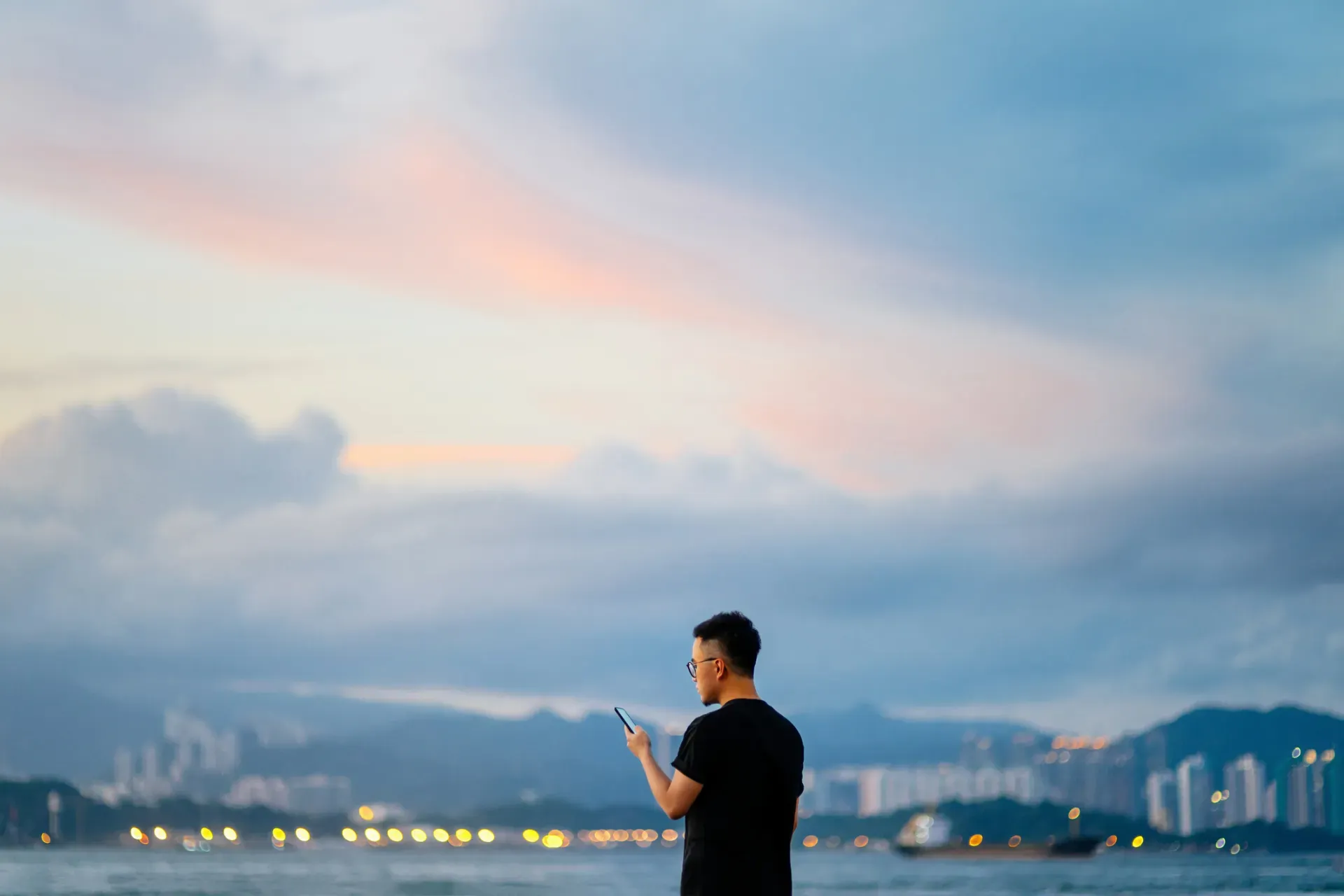 Person with phone silhouetted against twilight sky overlooking illuminated waterfront cityscape