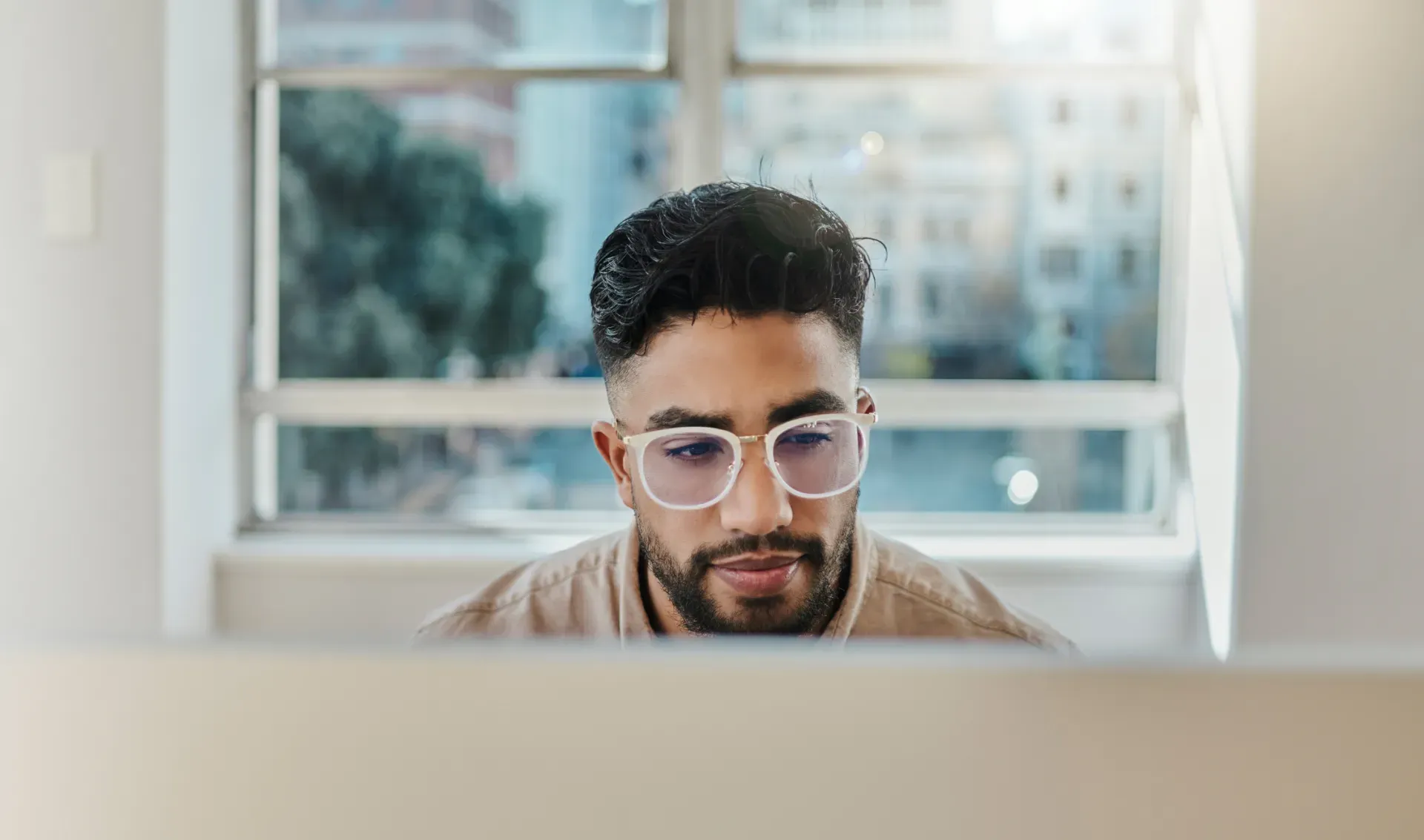 Young man with glasses focused on a computer screen in a bright office setting.