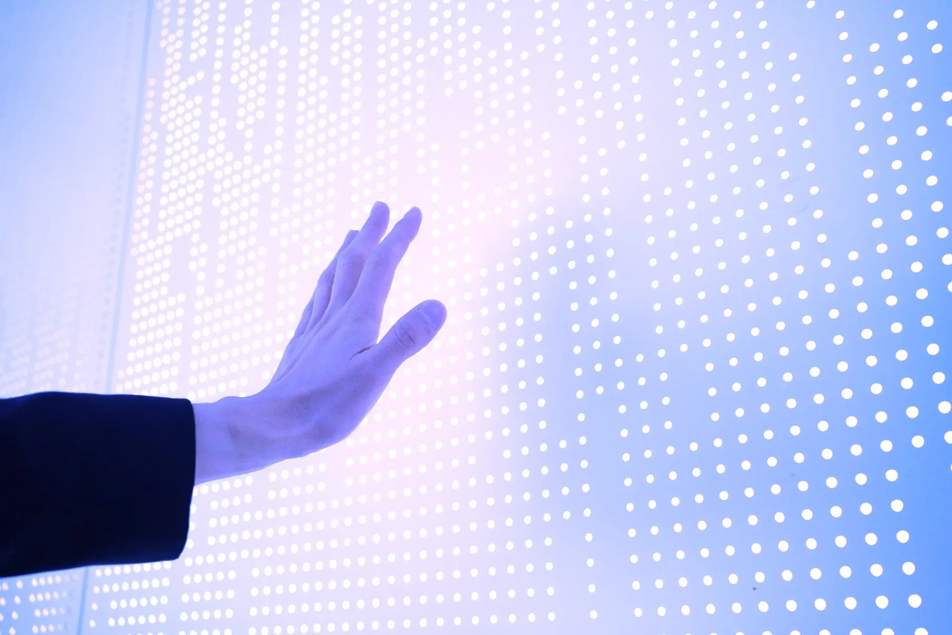 Hand reaching toward a glowing white LED dot-matrix panel with blue and purple light.
