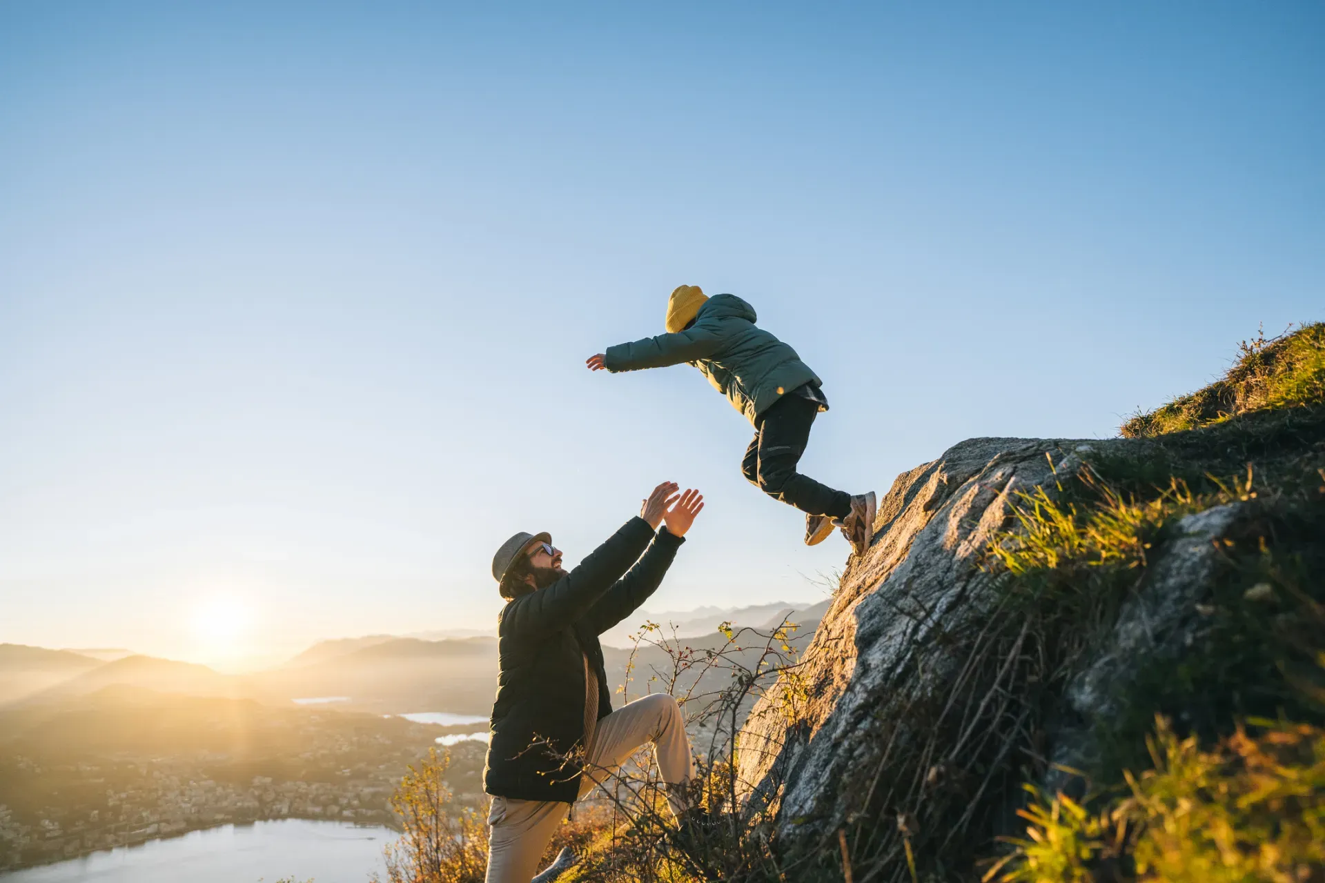 Adult lifting a child jumping off a rocky cliff at sunset, mountains in the background.