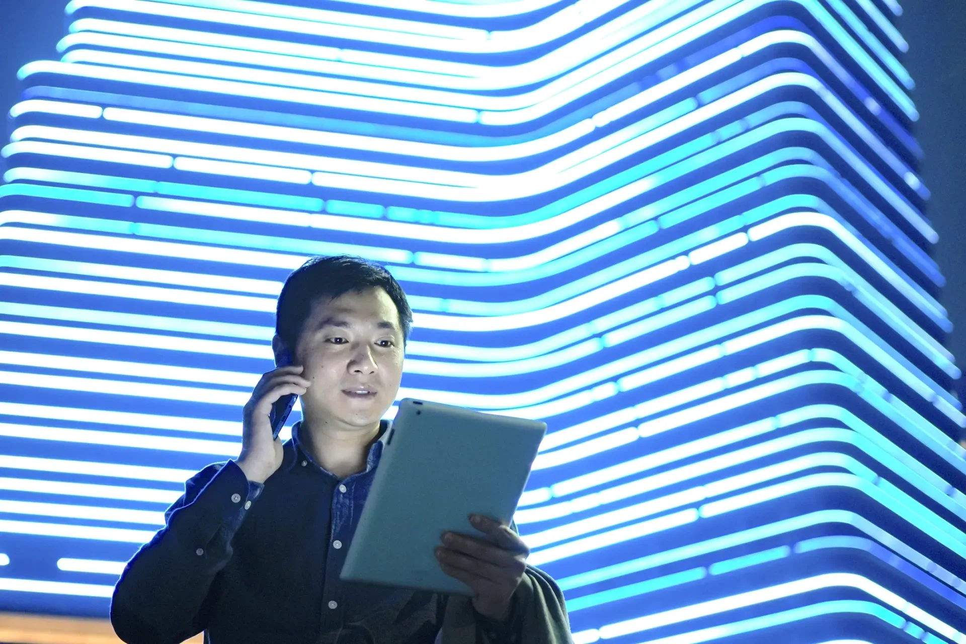 Man talking on phone while holding a tablet, standing in front of glowing blue neon lights. Man talking on phone while holding a tablet, standing in front of glowing blue neon lights.