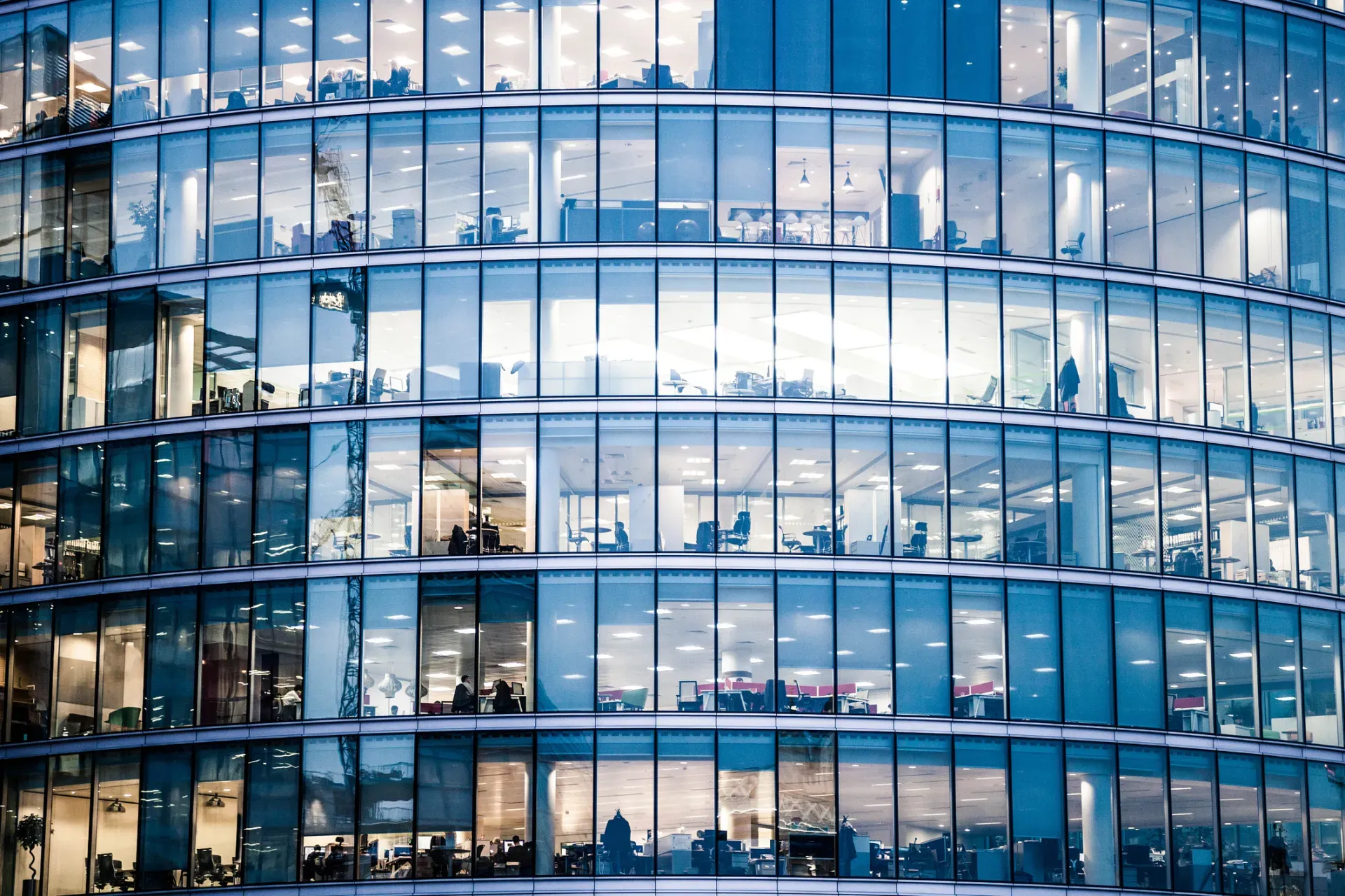 Modern glass office building facade with illuminated floors and people visible through windows. Modern glass office building facade with illuminated floors and people visible through windows.