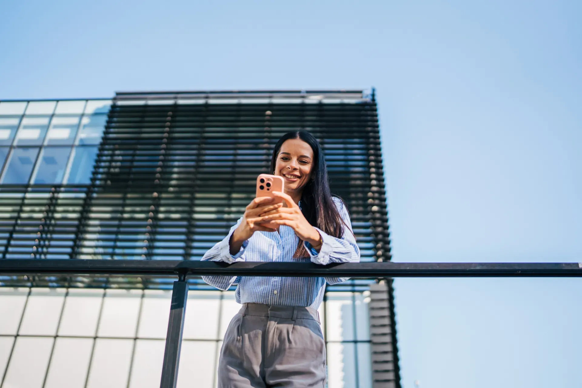 Young businesswoman smiling while using smartphone outdoors near a modern glass building. Young businesswoman smiling while using smartphone outdoors near a modern glass building.