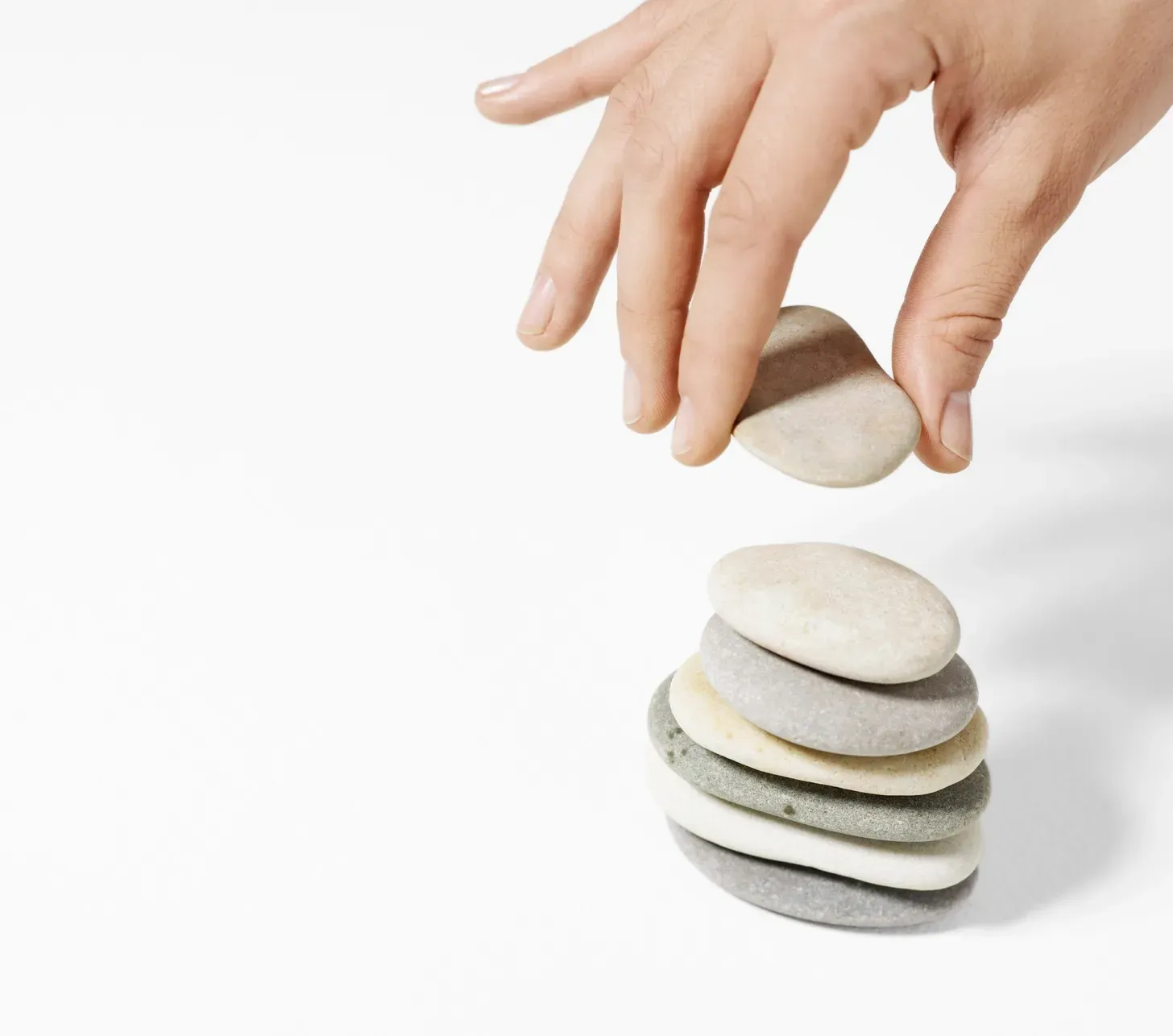 Hand placing a smooth stone on top of a balanced stack of pebbles against a white background. Hand placing a smooth stone on top of a balanced stack of pebbles against a white background.