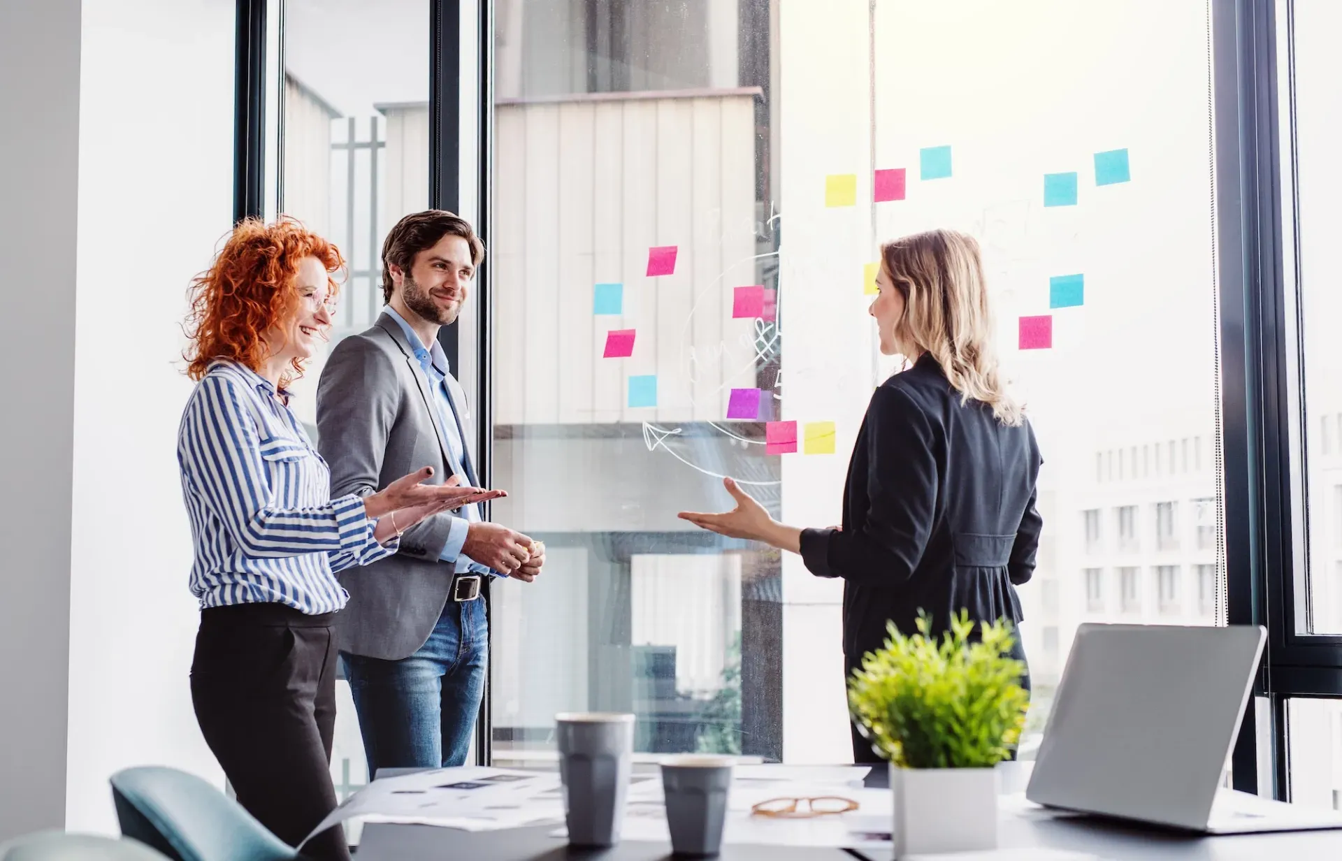 Three colleagues collaborating in a bright office, discussing colorful sticky notes on a glass wall. Three colleagues collaborating in a bright office, discussing colorful sticky notes on a glass wall.