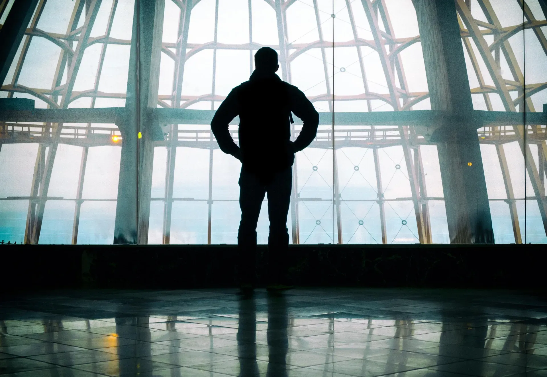 Silhouette of a person standing before large glass windows in a modern building, with light reflecting on the floor.